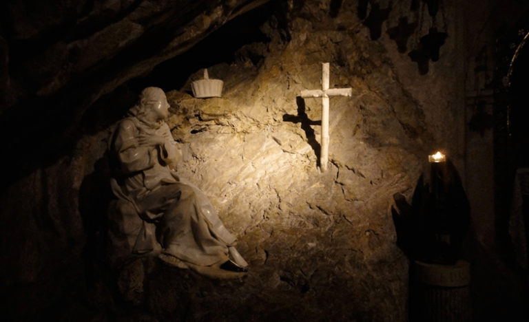 A statue of a saint gazes at a cross in a cave