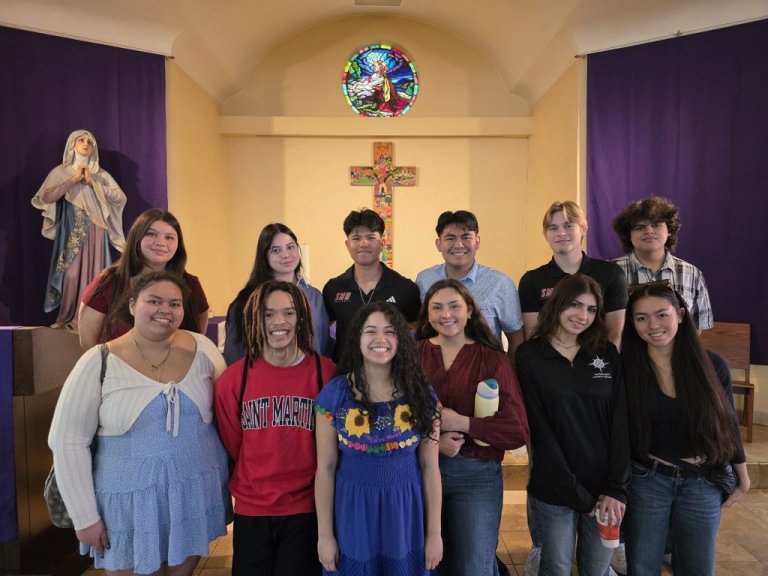 Students stand in a a chapel in front of a cross, statue of Mary, and stained glass