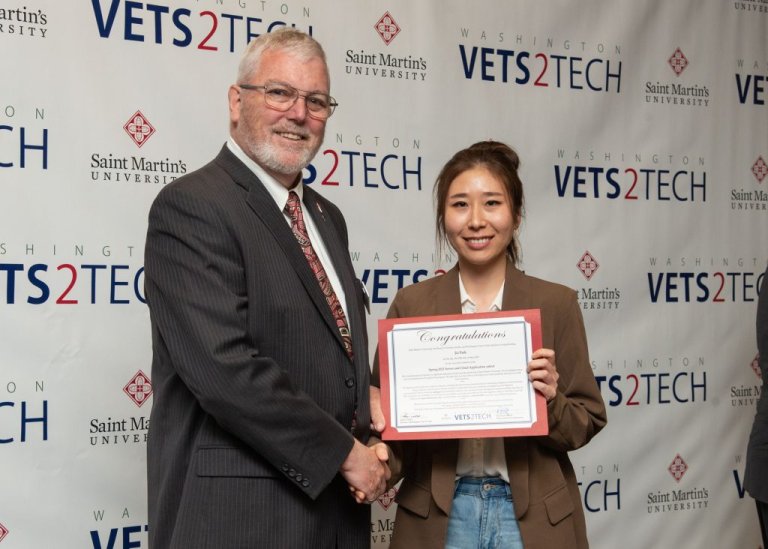 A student holds a certificate while shaking hands with an official in front of WaV2T backdrop