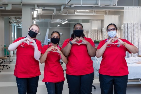 Four nursing students in scrubs and face masks pose making the Saints Care heart symbol with their hands.