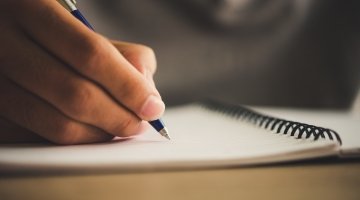 Close up of hand holding a pen writing in a spiralbound notebook.