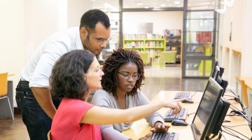 An instructor leans down to help two students working at a computer.