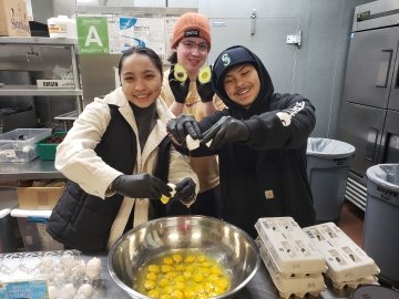 Students crack eggs at the community kitchen into a giant metal bowl full of eggs; one student holds up an avocado cut in half