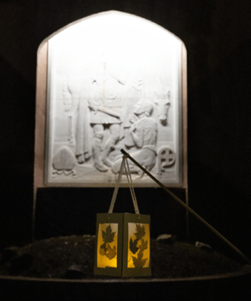 A lit, dangling handmade lantern with leaves in front of a carving at night