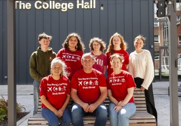 A group of students and faculty wearing red SMU study abroad shirts sitting on or standing around a bench